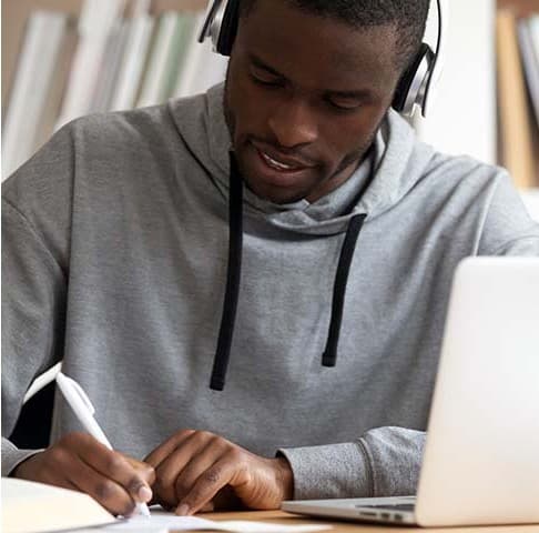 A student studying online with headphones, taking notes at a laptop